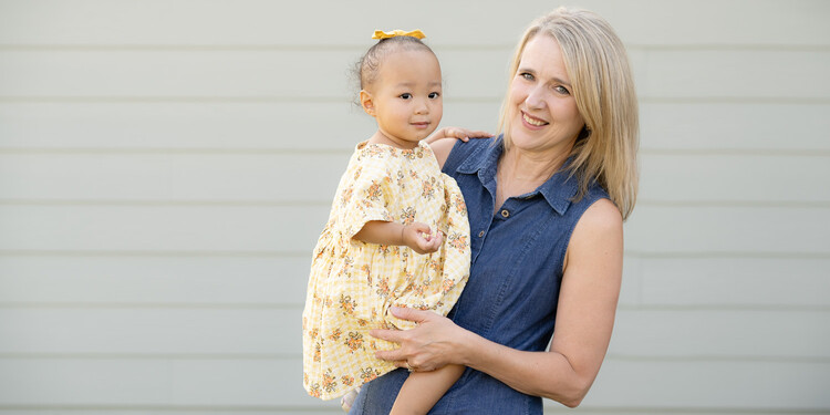 Darla holds Kara outside in front of the wall of a home