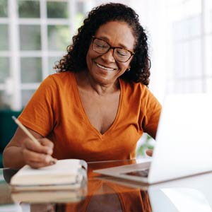 A woman smiles as she writers something in a notebook while working on a laptop